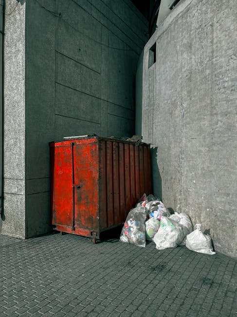 A red metal waste skip with a weathered, rusty surface positioned in a narrow alleyway at the junction of two modern building exteriors, one with dark green cladding and the other with a grey concrete finish. The skip is placed against the green wall on the left side, while several white plastic garbage bags, some tied and others loosely gathered, are stacked next to it on the textured dark paving surface. In the background, a small, rectangular window is visible on the grey wall, and part of a modern building's external corner extends vertically upward. The scene appears to depict private waste storage for rubbish collection, typical of independent waste disposal arrangements in an urban environment, often managed by local rubbish removal services such as those offered by Shepherds Bush specialists. The outdoor lighting is diffused, suggesting overcast conditions or shaded corridor access, with no natural vegetation or additional waste containers visible in this tight, utilitarian space.