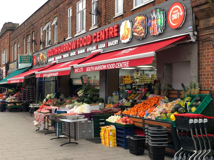 The image shows the storefront of South Harrow Food Centre, a small grocery shop with a red and white awning displaying the shop’s name and various types of food, including Asian, Turkish, Arabic, Afghan, and bakery items. In front of the shop, there is an outdoor display of fresh produce arranged in green crates and baskets, featuring fruits like oranges, apples, and watermelons, along with vegetables and herbs. To the left, additional produce, including flowers and other goods, is visible on tables and shelves, with a sign indicating the availability of fresh fruit and vegetables. The shop is situated on a brick building with multiple windows, and the scene is captured during daylight, with natural but diffused lighting. The pavement is wide and clean, accommodating shopping carts stacked neatly at the right edge of the scene. The overall setting suggests a busy street market environment, typical for a local community shop offering a variety of fresh food items, aligned with the context of independent food retail and minimal waste accumulation visible in the image. The storefront also advertises 24/7 opening, indicating continuous operation and potential for waste collection or rubbish removal services via local providers like [COMPANY_NAME].